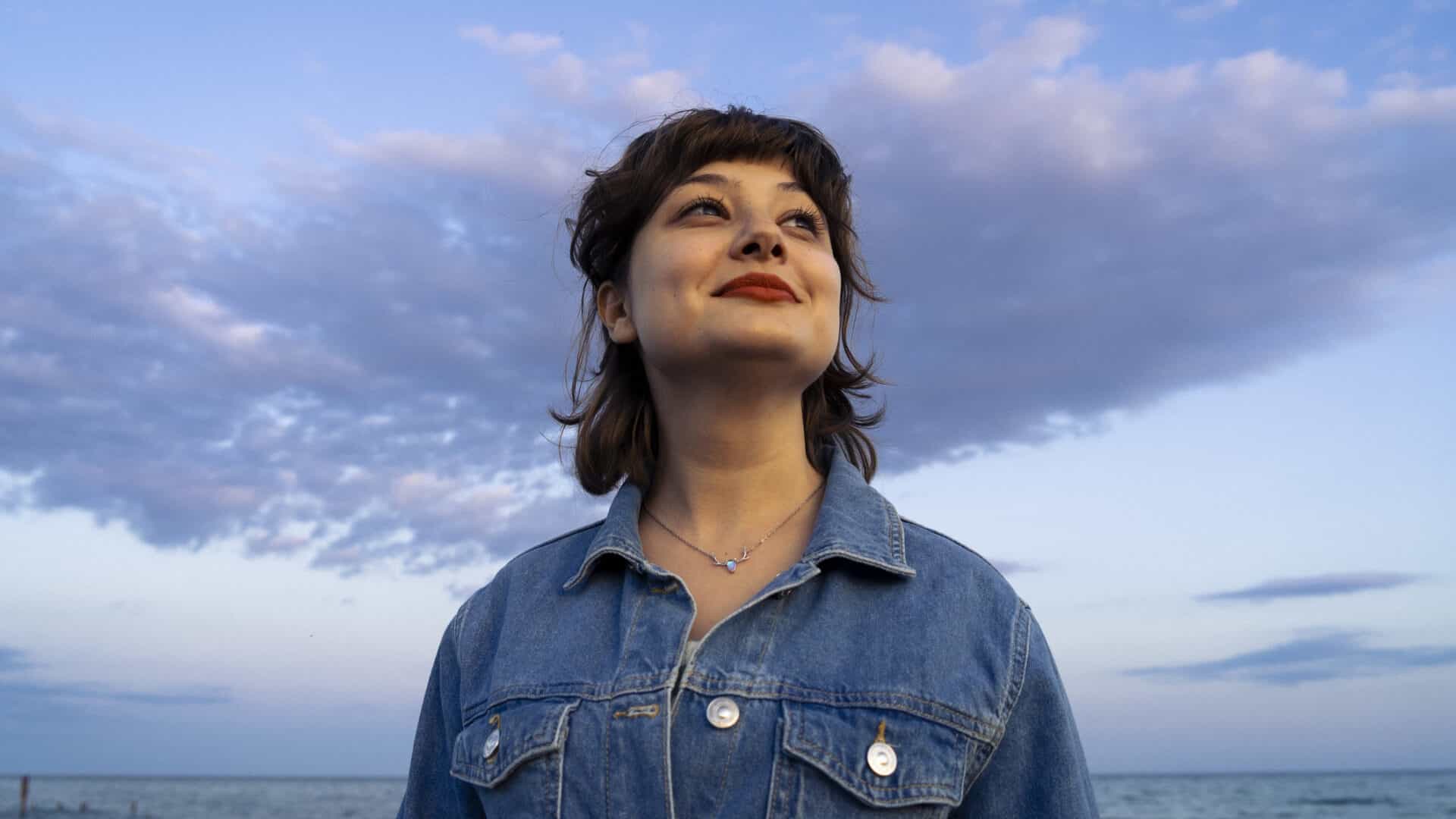 Photograph of a young woman wearing a denim jacket and smiling towards the sun, taken in the pastel colours of the sunset.